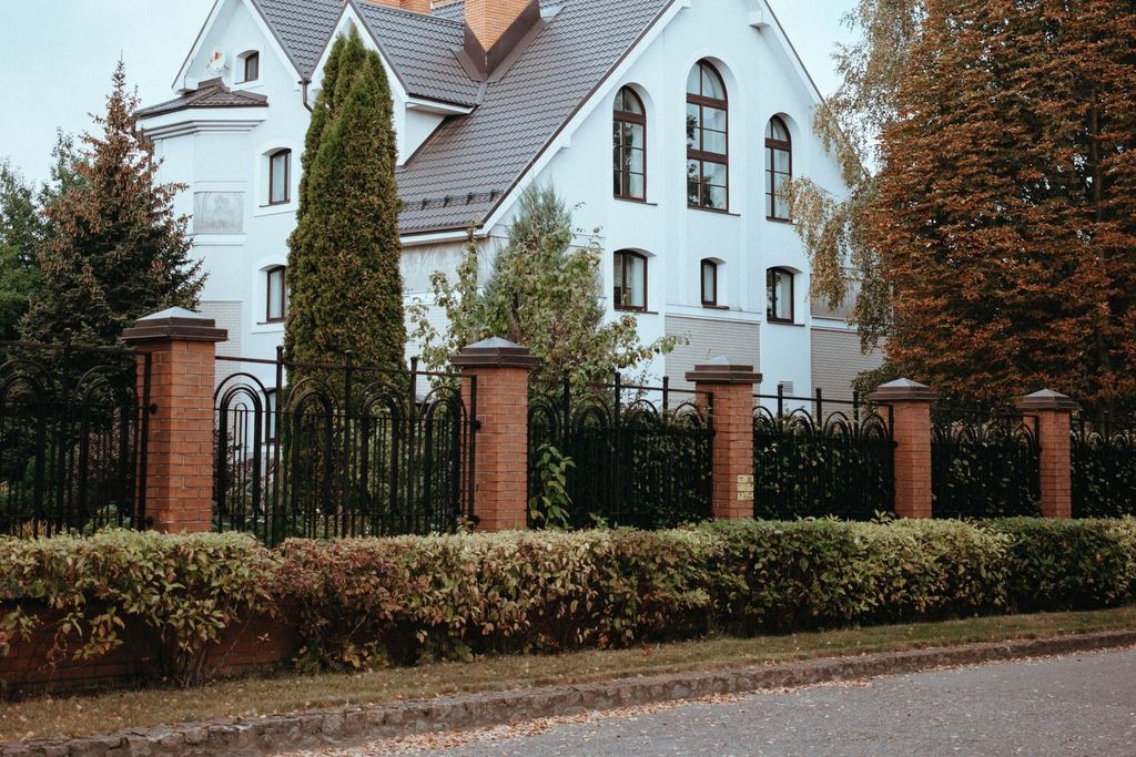 Vitt putsat stenhus med tegelpelare, smidesstaket och häck en höstdag