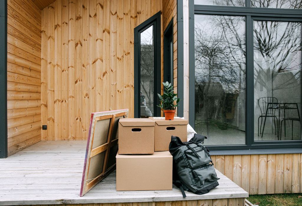 Flyttlådor, en ryggsäck, en krukväxt och en tavla på altanen framför ett nytt trähus.