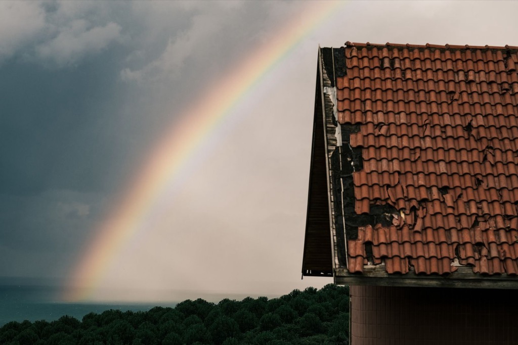 Slitet tegeltak med saknade pannor under en mörk himmel med regnbåge.